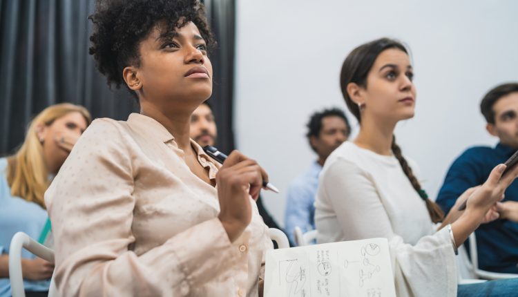 Foto de algumas pessoas assistindo a uma palestra. No primeiro plano, uma mulher preta, segurando uma caneta e um caderno, ao lado dela, em segundo plano, uma mulher morena, de cabelo liso e preso, no fundo, outras pessoas assistindo ao evento