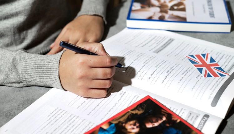 Foto focando em uma mão segurando uma caneta e escrevendo em um livro com vários exercícios e uma bandeira do Reino Unido, em cima de uma mesa ao sol