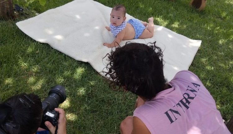 Fotógrafa tirando foto de bebê deitado em um cobertor esticado na grama, ao lado da profissional está a mãe do bebê, com uma camisa rosa escrita "interna"