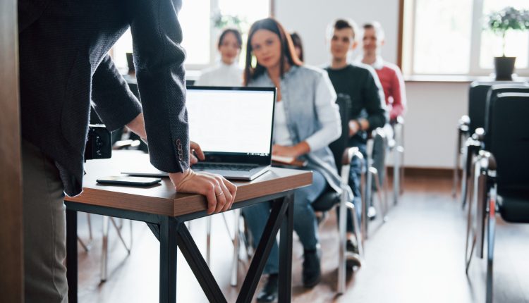 Um professor de costas, apoiando as mãos na mesa e ao fundo, uma sala de aula desfocada, com cadeiras enfileiradas e uma mulher sentada à frente e dois homens atrás