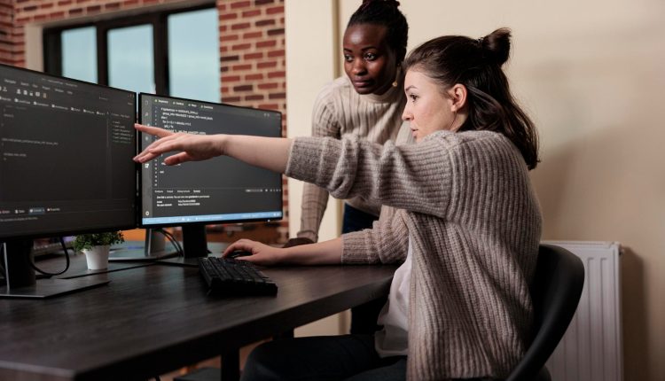 Duas mulheres em frente a duas telas de computador. Uma é asiática e está sentada apontando para a tela, a outra é uma mulher preta que está ao lado, olhando o que a outra mostra