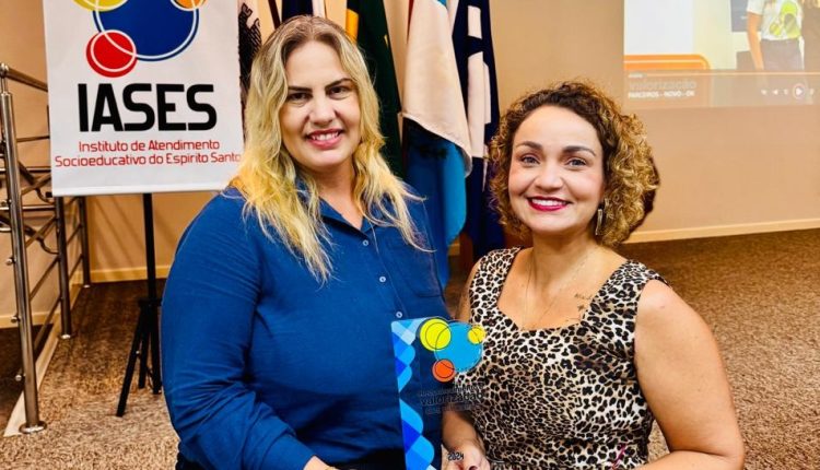 Duas mulheres, uma loira e alta e a outra morena de cabelo cacheado e mais baixa seguram um troféu nas mãos e posam para a câmera