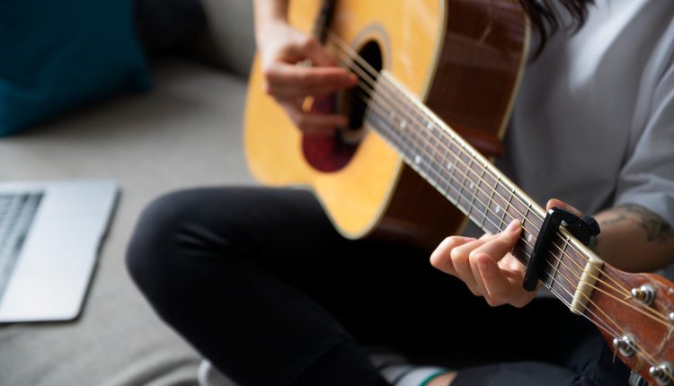 Foto do pescoço para baixo de uma pessoa tocando um violão, sentada e de pernas cruzadas em cima de um sofá