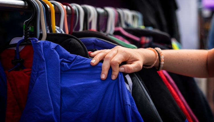 Uma arara com várias roupas penduradas por cabide e uma delas, uma jaqueta azul, sendo puxada para trás por uma mão branca