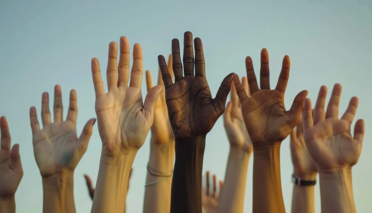 Foto de várias mãos de cores diferentes, levantadas, abertas e com as palmas viradas para a câmera